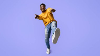 Sarı kazak giyen ve kot giyen genç bir adam, kamerayı işaret ederken geniş bir gülümsemeyle enerjik bir şekilde dans ediyor. Parlak mor arka plan, canlı sahneye eğlenceli bir dokunuş ekler..