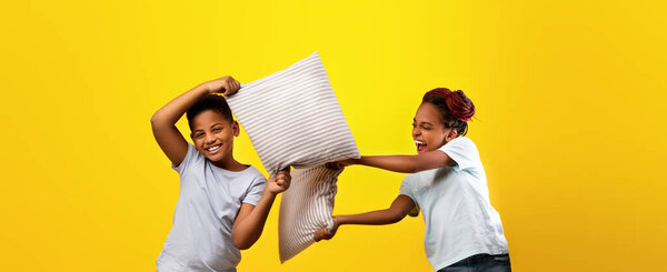 Two joyful children engage in a lively pillow fight against a vibrant yellow background. They smile and laugh, capturing the spirit of playful moments shared in childhood.