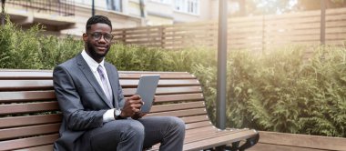 Takım elbiseli bir adam şehir parkındaki bir bankta rahatça oturur. Yeşillik ve modern mimari ile çevrili tabletine odaklanmış durumda. Aydınlık, güneşli bir gün..
