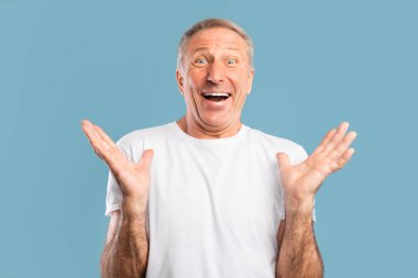 Beyaz tişörtlü neşeli olgun bir adam kameraya geniş gözlü ve ağzı açık bakar. Mavi bir stüdyo arkaplanına karşı coşkuyla ellerini kaldırır. Saf bir zevk anı yakalar..