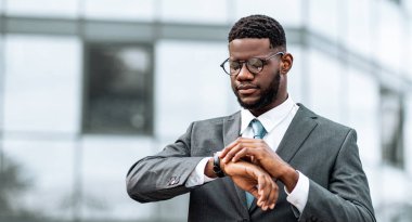 Takım elbiseli bir adam çağdaş bir ofis binasının önünde durmuş saatine odaklanmış bir ifadeyle bakıyor. Gözlük takıyor ve önemli bir toplantı ya da etkinlik bekliyor gibi görünüyor..