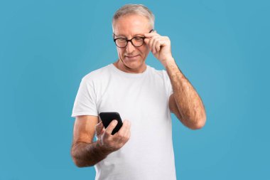 Mutlu, olgun bir adam mavi bir stüdyoda duruyor ve akıllı telefonuyla meşgul. Sosyal medyada gezinirken, gözlüğünü düzeltirken ve neşeli görünürken gülümsüyor..