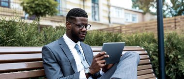 Takım elbiseli bir adam parkta bir bankta oturuyor, tabletine odaklanmış. Gözlük takıyor ve gün boyunca çevresi yeşillik ve kentsel binalarla çevrili olarak işine dalmış gibi görünüyor..