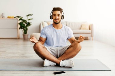 Genç bir adam parlak bir oturma odasında yoga minderinde bacak bacak üstüne atmış oturuyor. O, meditasyon uygulamasına odaklanırken kulaklıkla sakinleştirici müzik dinler ve huzurlu bir atmosferin tadını çıkarır..
