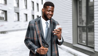 Şık bir ekose ceket giymiş ve akıllı telefonuna bakarken gülümseyen bir adam. Diğer elinde bir kahve fincanı tutuyor ve etrafındaki binalarla modern bir cadde boyunca yürüyor..