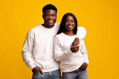 Genç bir çift birbirine yakın duruyor, ikisi de beyaz kazak ve rahat kot pantolon giyiyor. Neşeli bir şekilde gülümsüyorlar, mutluluklarını canlı sarı bir zemin önünde sergiliyorlar..