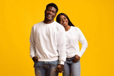 Mutlu bir çift birbirine yakın duruyor, ikisi de beyaz kazak giyiyor. Parlak sarı bir arkaplana gülümsüyorlar, neşeli ve rahat bir ortam sergiliyorlar..