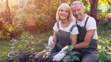 Neşeli bir çift arka bahçelerinde bahçeyle eğleniyor. Onlar canlı bitkiler ve güneş ışığıyla çevrili çimlerin üzerinde otururken çiçek ekiyorlar. Gülümsemeler bu aktivitedeki neşelerini yansıtıyor..
