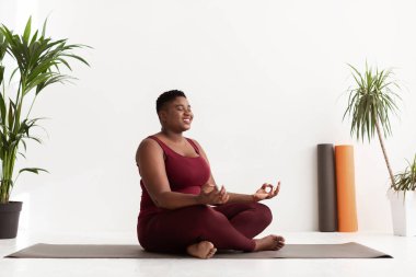 Kısa saçlı şişman siyah kadın nilüfer pozisyonunda bir spor minderinin üzerinde oturur, sakin bir yoga merkezinde gözleri kapalı meditasyon yapar. Çevresini çevreleyen yeşil bitkiler, sakin bir atmosfer yaratıyor..