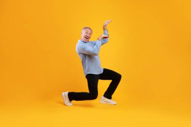 Neşeli, olgun bir adam, canlı sarı bir arka planda müzik eşliğinde dans ederken gülümser. Heyecanını bu heyecanlı anda eğlenceli disko hareketleriyle ifade ediyor..