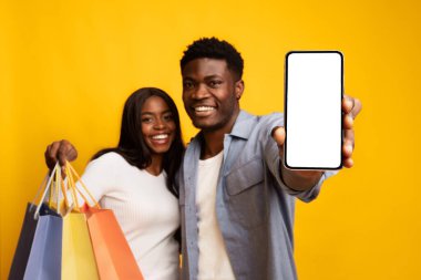 Genç bir çift yan yana durur, gülümser ve alışveriş torbalarını tutarken adam gururla boş ekranlı akıllı telefonu gösterir. Parlak sarı bir arka planın önündeler..
