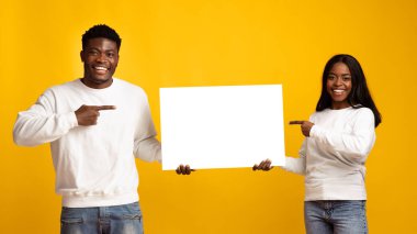 Neşeli genç bir adam ve kadın yan yana duruyor, geniş bir gülümsemeyle. Her biri ellerinde tuttukları boş bir tabelayı işaret ediyor, neşeli tavırlarını artıran canlı sarı bir zemin ile..
