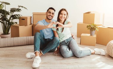 Mutlu bir çift, etrafı kutularla çevrili aydınlık bir odada yerde oturuyor. Elleriyle yeni başlangıçlarını kutlamak için kalp şekli oluşturuyorlar..