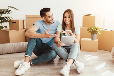 Bir çift, kutuları taşıyan, gülümseyen ve sohbete dalmış bir şekilde yerde oturuyor. Yeni alanlarını düzenlemek için atacakları sonraki adımları planlarken ellerinde bir tablet tutuyorlar..