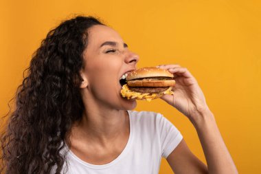 Kıvırcık saçlı neşeli bir kadın büyük bir hamburgeri ısırıp abur cubura olan aşkını gösteriyor. Canlı sarı bir arkaplanda poz veriyor, oburluğu neşeli bir şekilde kutluyor..