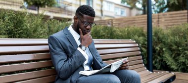 Gri takım elbiseli bir adam parkta bir bankta oturuyor ve gazete okumaya odaklanıyor. Uzun otlar ve ağaçlar güneşli bir öğleden sonra dingin bir zemin sağlar..