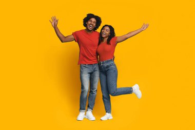 Neşeli bir çift, kollarını heyecanla kaldırarak, geniş gülücüklerle bir arada durur. Parlak sarı bir duvara karşı şakacı poz verirken kırmızı bluz ve kot pantolon giyerler..