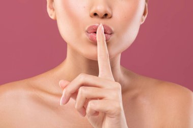 Bir kadın yakın poz verir ve sessiz bir jest yapar. Parmağı dudaklarındadır. Dolgun dudakları var ve mor arka plan çekim için yumuşak bir ortam yaratıyor..