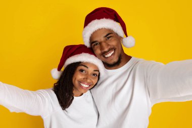 Mutlu bir çift, her ikisi de kırmızı Noel Baba şapkası takan bir selfie için poz veriyor. Işıldayan gülümsemeleri, bayram neşesini canlı sarı bir arka plana yayarak mevsimin şenlikli ruhunu yakalıyor..