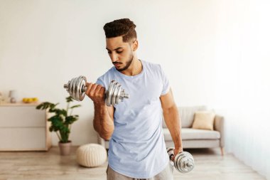 İyi aydınlatılmış bir odada egzersiz yapan bir adam, odaklanmış bir kararlılıkla bir dambılı kaldırır. Mekan kapalı bitkilerle dekore edilmiş ve rahat mobilyalar içeriyor..