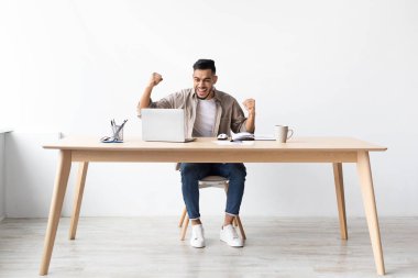 Sakallı neşeli bir adam ev ofisinde çalışırken bir başarıyı kutluyor. Tahta bir masada oturuyor, heyecan içinde yumruklarını kaldırıyor ve dizüstü bilgisayarına gülümsüyor..