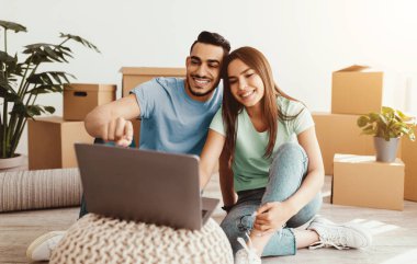 Bir çift, kutuları taşırken dizüstü bilgisayarla mutlu mesut yerde oturuyor. Gülümsemeleri ve kahkahaları paylaşıyorlar, yeni alanlarında eğleniyorlar, birlikte anılar yaratıyorlar..