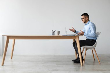 Arap bir adam ofisinden bir video görüşmesi yapıyor. O bir masada oturur, dizüstü bilgisayarla konuşurken anlamlı bir şekilde el kol hareketi yapar. Ayar parlak ve profesyonelce..