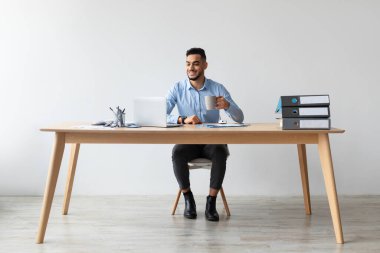 Neşeli bir Arap, çağdaş bir ofiste minimalist bir masada oturur ve bir fincan kahvenin tadını çıkarırken dizüstü bilgisayarını kullanır. Odaklanmış ve memnun görünüyor, verimli bir çalışma ortamı sergiliyor..
