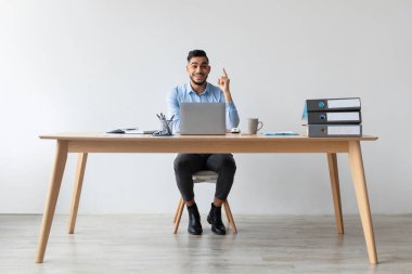 Genç iş adamı bir dizüstü bilgisayarla şık bir masada oturuyor, gülümsüyor ve bir anda parmağını kaldırıyor. Parlak bir ofis ortamında heyecan ve yaratıcılığı somutlaştırıyor..