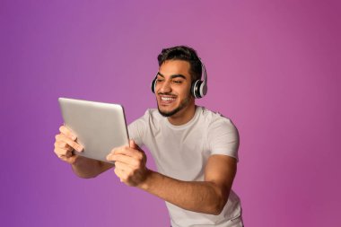 Genç bir Arap adam kulaklık takarken tablet ekrana bakarak eğleniyor. Modern neon ışıkları, cihazındaki bir uygulamayla bağlantı kurarken canlı bir atmosfer yaratıyor..