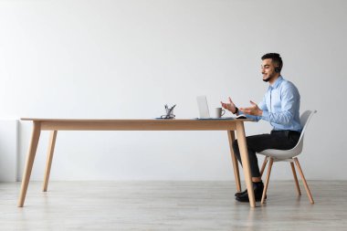 Arap bir adam geniş bir ofis masasında oturuyor, kulaklık takıyor ve bilgisayarında video görüşmesi yapıyor. Müşterilerle uzaktan iletişim kurarken gülümsüyor, işinden zevk alıyor..