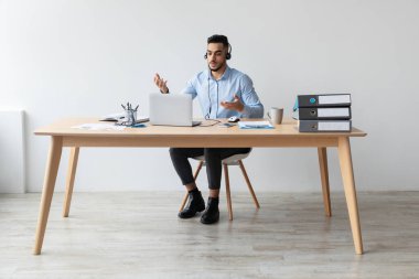Arap bir adam ofis masasında otururken bir video görüşmesinde müşterileriyle dizüstü bilgisayarla iletişim kuruyor. Profesyonel olarak giyinmiş, online toplantı sırasında net ses için kulaklık takıyor..