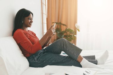 Genç bir kadın, akıllı telefonuyla meşgul bir şekilde kanepede rahatça oturur. Güneş ışığı pencereden akar ve sıcak bir atmosfer yaratır..