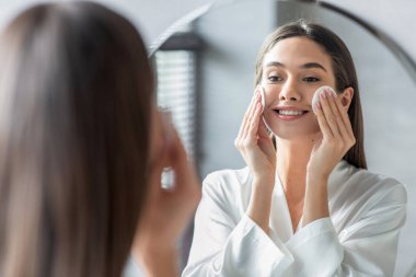 Genç bir kadının pamuk pedlerle cildini temizlediği parlak bir banyo sahnesi. Bir aynanın karşısında duruyor, mutlu görünüyor ve günlük cilt bakımı sırasında yansımasına odaklanıyor..