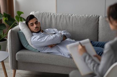 Genç bir Arap adam terapi ofisindeki bir kanepede uzanmış stres ve depresyon duygularını tartışıyor. Psikologu seans sırasında dikkatle dinlerken notlar alıyor..