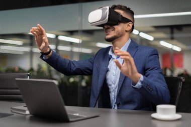 Heyecanlı iş adamı çalışma masasında oturur, VR gözlük takar ve havaya dokunur. Çağdaş bir ofis ortamında modern teknolojiyi sergileyen sanal elementlerle uğraşır..