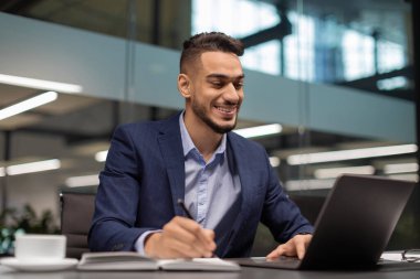 Takım elbiseli neşeli bir Arap girişimci şık bir ofisteki dizüstü bilgisayarına odaklanır. Yeni iş fırsatlarını araştırırken, iş ortamının tadını çıkarırken notlar alıyor..