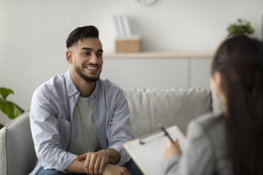 Orta Doğulu bir adam ruh sağlığı kliniğindeki bir kanepede rahatça oturup psikoloğuyla konuşurken gülümsüyor. Seans boyunca coşku ve açıklık göstererek terapi sonuçlarını paylaşıyor..