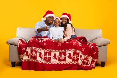 Üç kişilik aile, kırmızı bir battaniyeyle kaplı bir kanepede oturur ve Noel film gecesinin tadını çıkarırlar. Sıcak sarı duvar Noel ruhunu artırırken onlar şenlikli şapkalar giyerler..