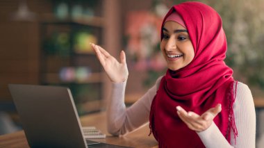 Kırmızı tesettürlü genç bir kadın bir video çağrısına katılıyor, gülümsüyor ve hareketli bir şekilde işaret ediyor. Sıcak, davetkar bir yerde gibi görünüyor. Arka planda hafif ışıklandırma ve yeşillik var..