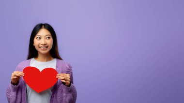 Genç bir kadın, önünde kocaman kırmızı bir kalp şeklini tutarken gülümsüyor. Arkaplan, onun neşesini vurgulayan neşeli bir atmosfer yaratan katı mor bir renk..