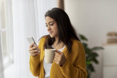 Genç Arap bayan evde pencerenin kenarında oturuyor, akıllı telefonunu karıştırırken bir fincandan kahve yudumluyor. Arkadaşlarıyla mesajlaşıyor, rahatlatıcı bir anın tadını çıkarıyor..