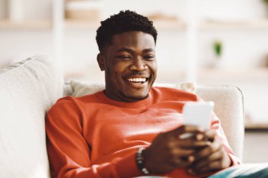 Neşeli genç bir adam bir kanepede rahat bir şekilde oturur, akıllı bir telefon tutar ve gülümser. Doğal ışık odayı doldurur, sıcak ve davetkar bir atmosfer yaratır..