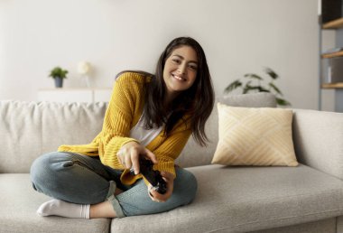 Ortadoğulu mutlu bir kadın rahat bir oturma odasında koltuğunda oturup oyun oynayarak eğleniyor. Evde kaldığı süre zarfında rahatlamış ve eğleniyor..