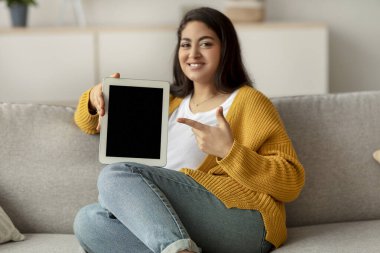 Neşeli bir Arap kadın bir kanepede oturup tablet ekranı işaret ediyor. İnternetten alışveriş yapmayı ve evde hediye aramaktan hoşlanıyor. Ortam rahat ve davetkar, dijital alımlar için mükemmel..