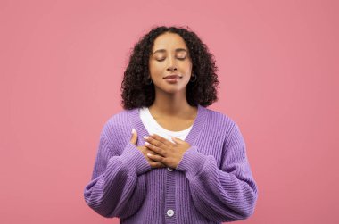 Samimi, genç, siyah bir kadın elleri kalbinin üstünde, gözleri kapalı ve huzurlu bir minnettarlık ifadesi içinde duruyor. Yumuşak pembe bir arka planı olan bir stüdyoda sıcaklığını ve takdirini iletiyor..