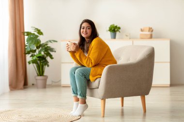 Genç bir Arap bayan koltukta rahatça otururken kameraya gülümsüyor. Bir fincan sıcak kahve tutuyor, serin bir günde sıcak içeceğin tadını çıkarıyor, sıcak bir atmosfer yaratıyor..