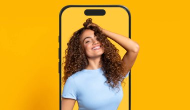 Kıvırcık saçlı, gülümseyen ve neşeli bir poz veren bir kadın akıllı telefon çerçeveli, canlı sarı bir arka plana karşı. O anda neşe ve güven yayar..