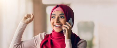 Parlak kırmızı tesettürlü genç bir kadın heyecanla gülümser ve elini kaldırır. Işıkla dolu, rahat bir yaşam alanında telefonla konuşurken..