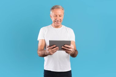 Kendine güvenen olgun bir adam mavi bir duvarın önünde elinde tablet tutarken parlak bir şekilde gülümsüyor. İçeriği izlemekle meşgul ve neşeli ve pozitif görünüyor..